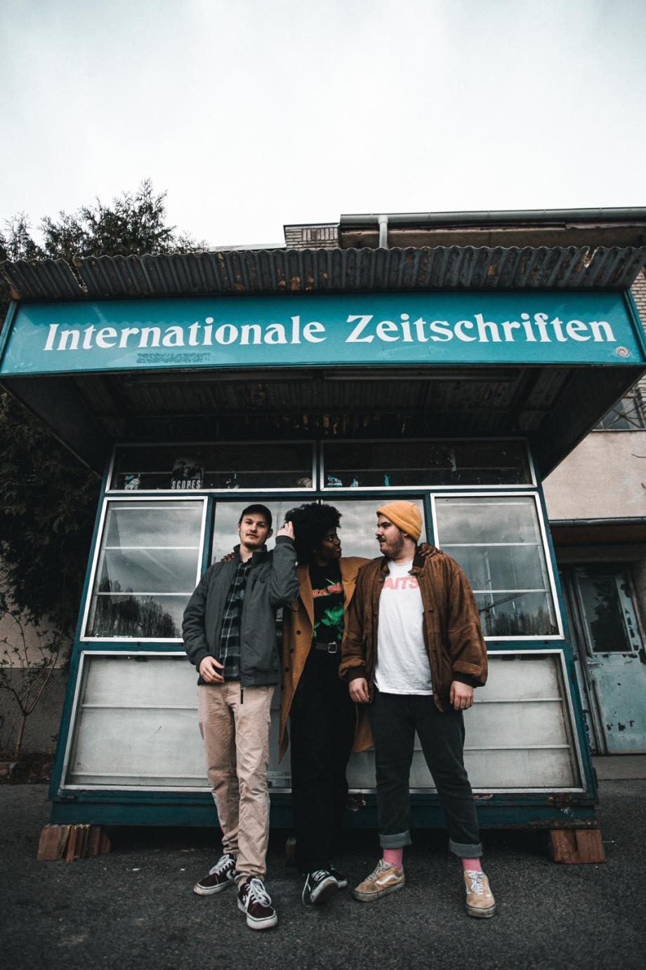 Die drei Mitglieder von Curb stehen vor einem Zeitungsstand mit der Aufschrift "Internationale Zeitschriften""