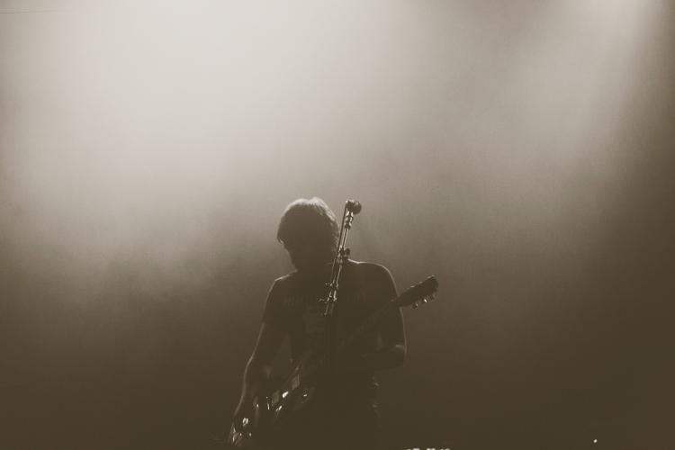 Die Silhouette eines Musikers mit Gitarre, vor ihm steht ein Mikrofonständer, das Bild ist dunkel gehalten.